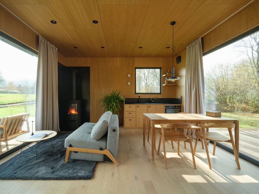 Holzverkleideter Wohnraum einer Dwellii-Kabine: Holzofen, graues Sofa, Essgruppe aus Eiche, zwei raumhohe Verglasungen zur Wiese