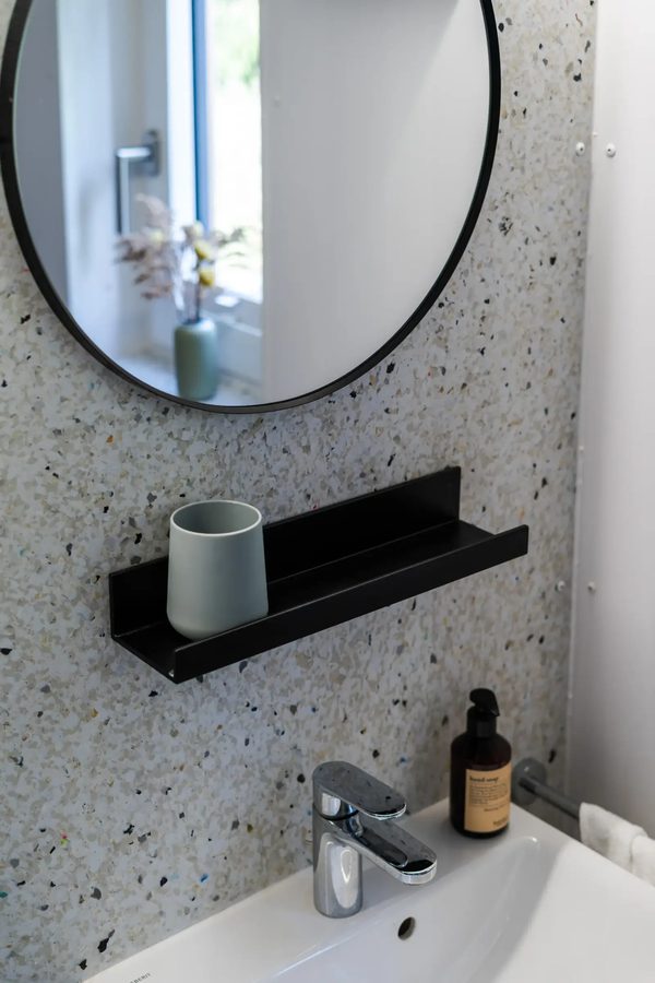 Bad der Tinca Cabin im Detail: weißes Waschbecken, runder schwarzer Spiegel, Wand in Terrazzo-Optik, schwarzes Regal