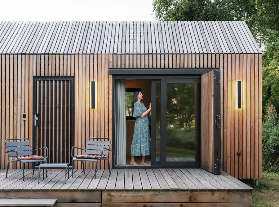 Fassade der Tinca Cabin im Detail: Lärchenlamellen, geöffnete Schiebetür mit schwarzem Rahmen, Terrassenstühle, eine Bewohnerin auf dem Deck