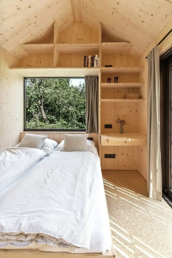 Schlafbereich der Tinca Cabin: weiße Bettwäsche, sichtbare Holzdecke im Giebel, Leiterregal und Fenster ins Grüne