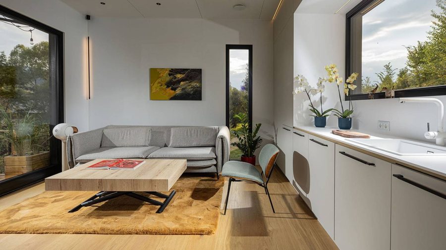 Wohnbereich des TR01 mit Küchenzeile: graues Sofa, Couchtisch, weiße Fronten, Fenster auf beiden Seiten