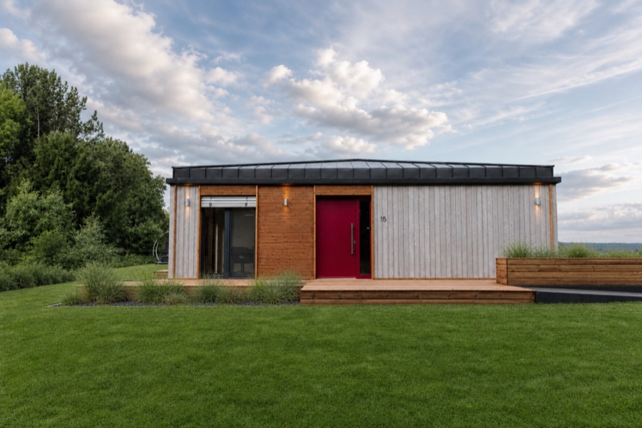 Gartenfassade des Musterhauses TR3M auf weiter Rasenfläche, rote Eingangstür, großer Himmel mit Wolken
