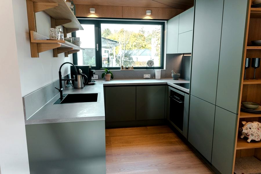 Küche im Musterhaus TR3M in Salbeigrün: Fenster über der Spüle, Einbaubackofen, offene Eichenregale, Steinarbeitsplatte