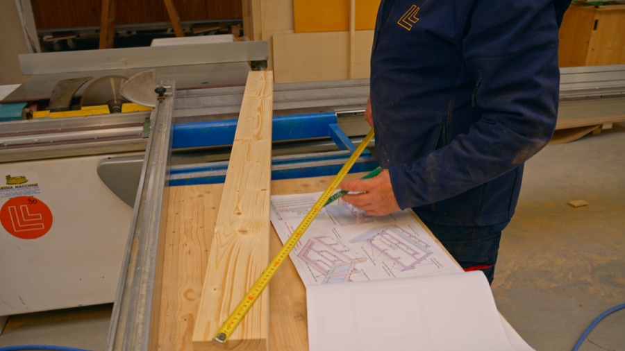 Mitarbeiter gleicht Bauplan, Maßband und Holzbauteil vor dem Zuschnitt ab